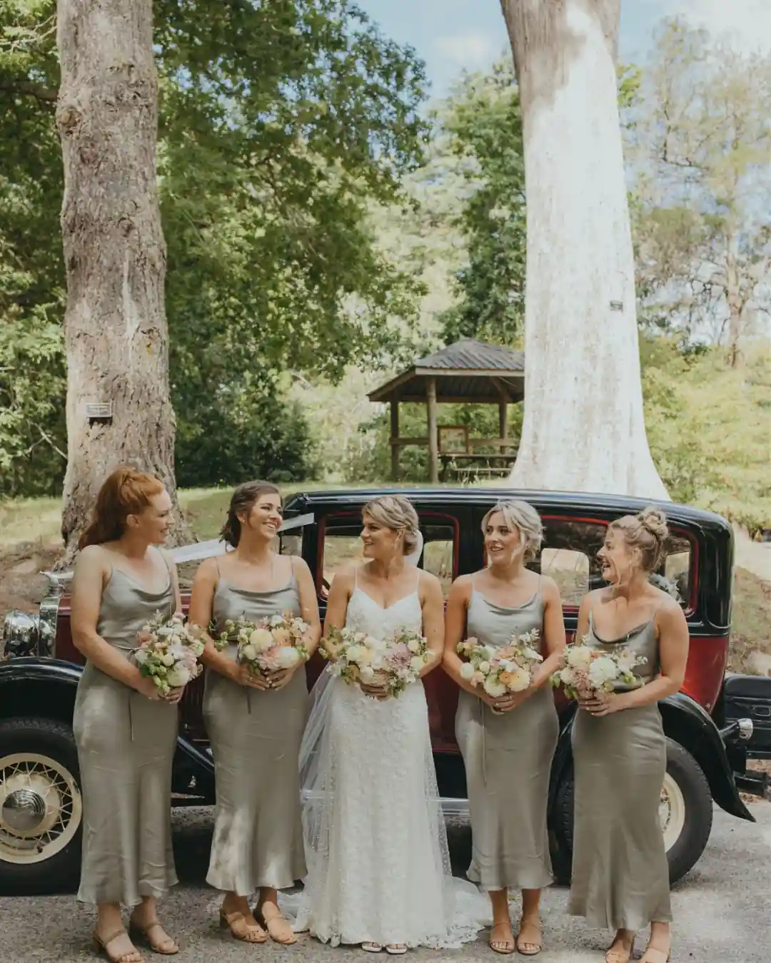 Bridesmaids in sage dresses beside vintage car at arboretum