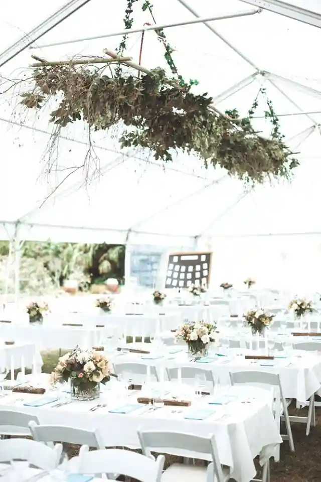 Gisborne arboretum wedding marquee setup with white tables, flower centrepieces and hanging branch greenery